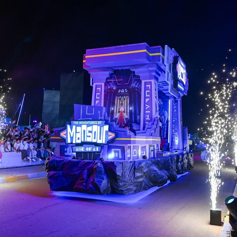 Illuminated purple Mansour Festival gateway float moving through the parade at night beside pyrotechnic effects.